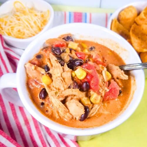 Chicken chili in a white bowl with cheese and corn chips in small bowls. Red and white dish towel.