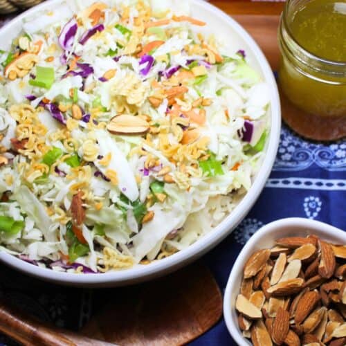 Ramen noodle coleslaw on a table with a blue cloth, dressing, and nuts.