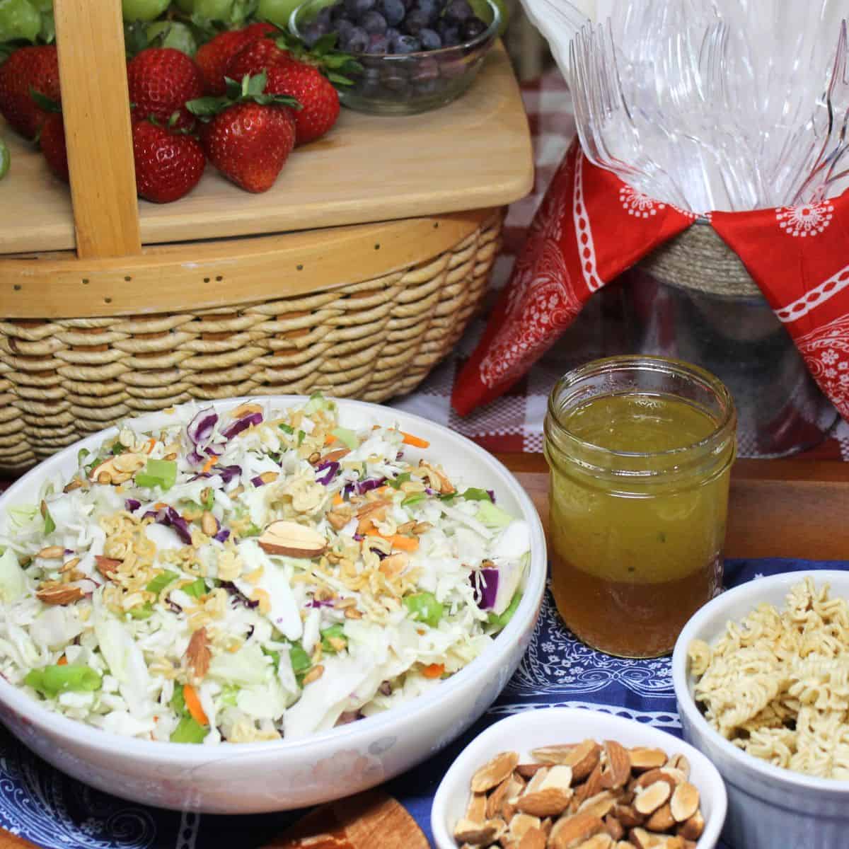 Ramen Noodle Coleslaw in a bowl with a basket, fruit, forks, dressing, almonds, and toasted noodles in the picture.